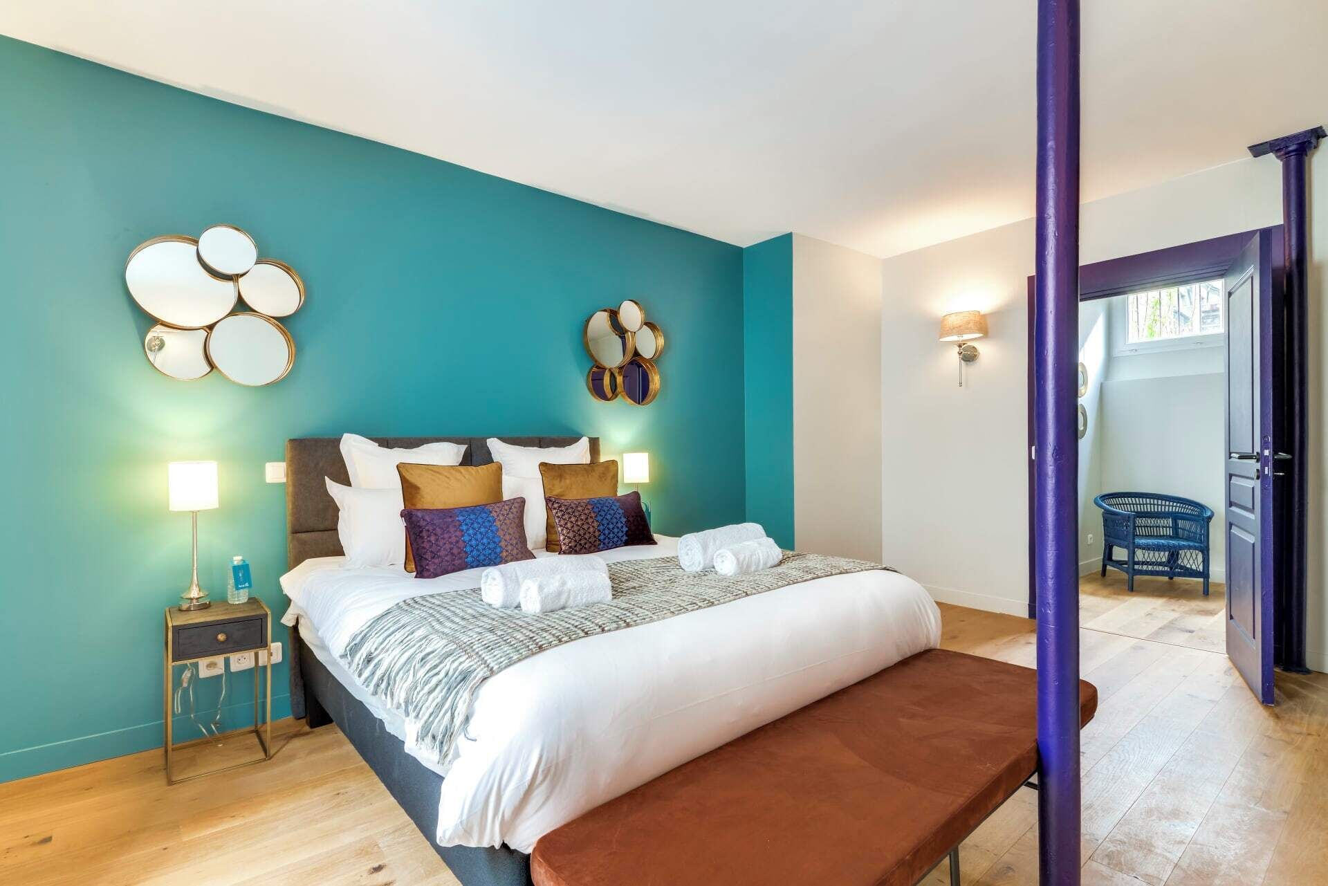 A modern bedroom with a teal wall, a large bed with multiple pillows, a bedside table with a lamp, and a bench.