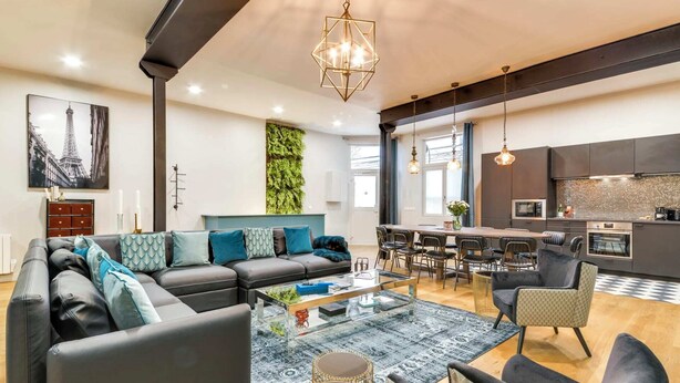 A modern living room with a sofa, coffee table, and chairs. There is a dining area with a table and chairs. A kitchen is visible with modern appliances.