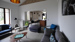 Un salon moderne avec un canapé, un pouf et une table basse. On y trouve également un coin repas avec une table et des chaises, et une cuisine visible au fond.