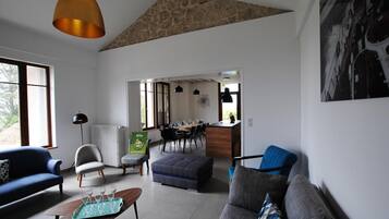 Un salon moderne avec un canapé, un pouf et une table basse. On y trouve également un coin repas avec une table et des chaises, et une cuisine visible au fond.