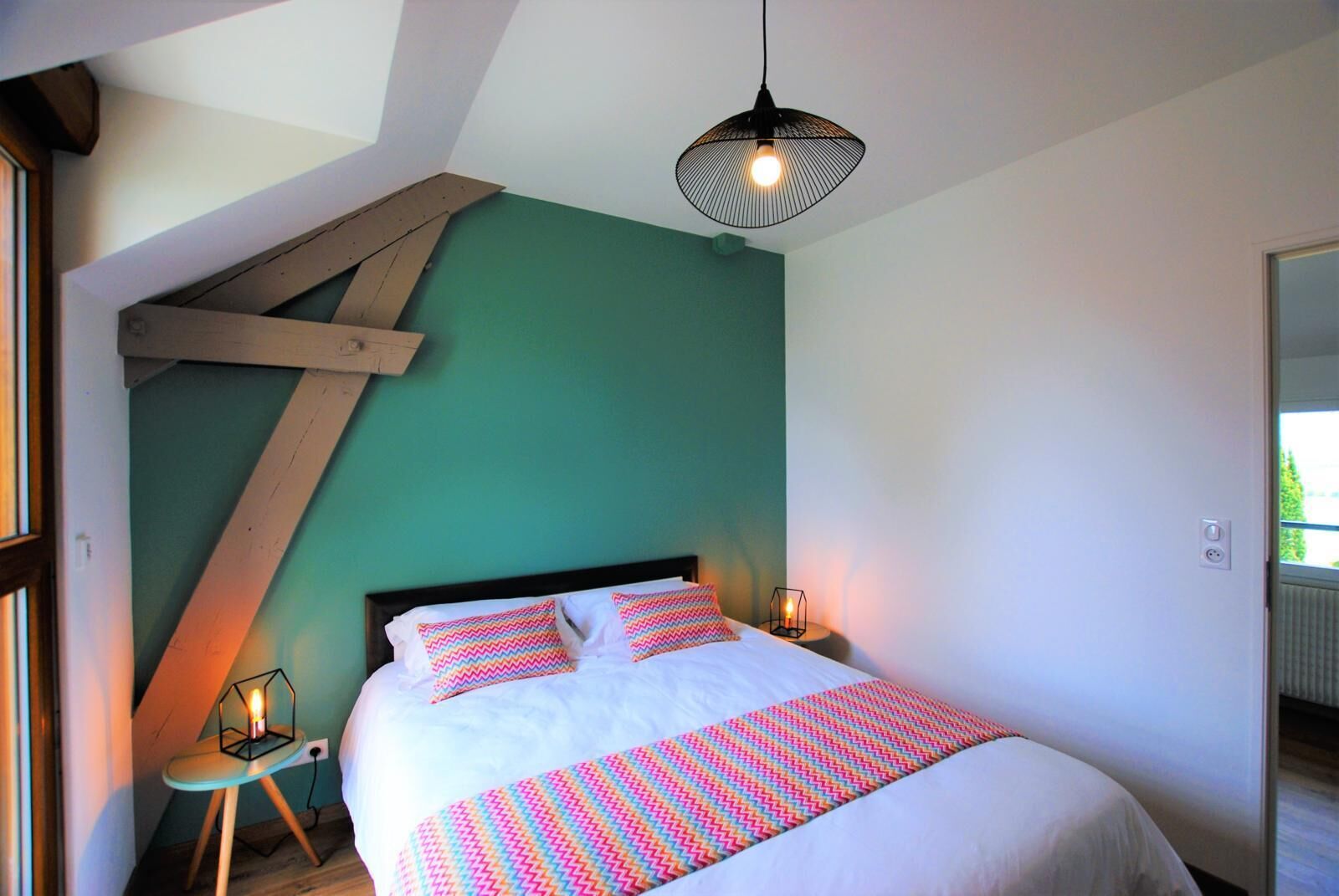 Een slaapkamer met een schuine kap, een bed met witte lakens en kleurrijke kussens, een hanglamp en een klein tafeltje met een kaars.
