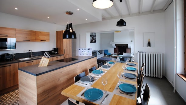 Une cuisine moderne avec des meubles en bois, un îlot central et un coin repas aménagé pour six personnes, avec de l’assiette et des verres bleus.