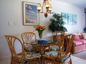 Un rincón comedor con una mesa redonda de vidrio, sillas de madera con tapicería floral, un jarrón con flores y una lámpara fijada a la pared.