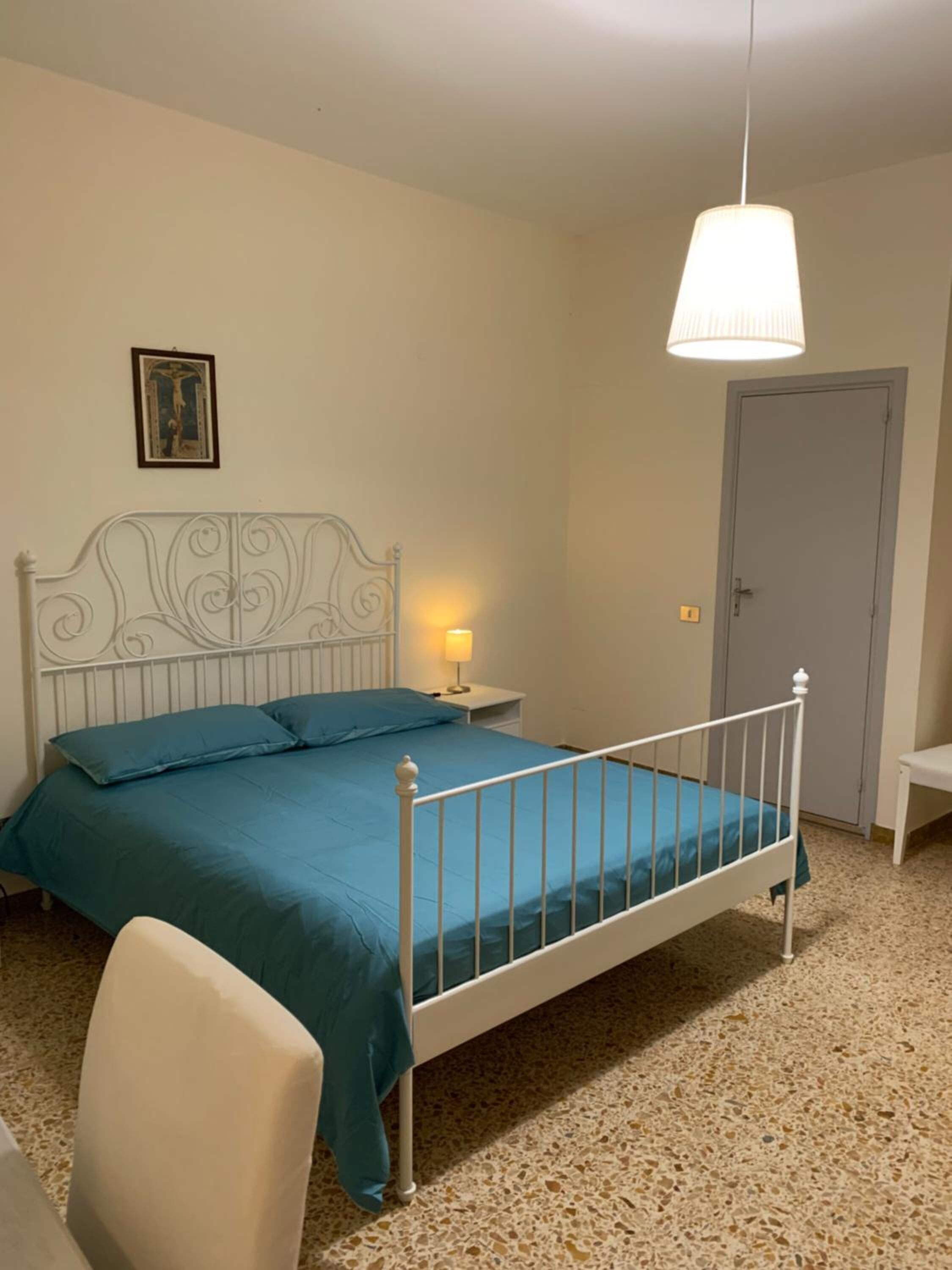 A bedroom with a white metal bed frame, teal bedding, a white nightstand with a lamp, a framed picture on the wall, and a closed door.