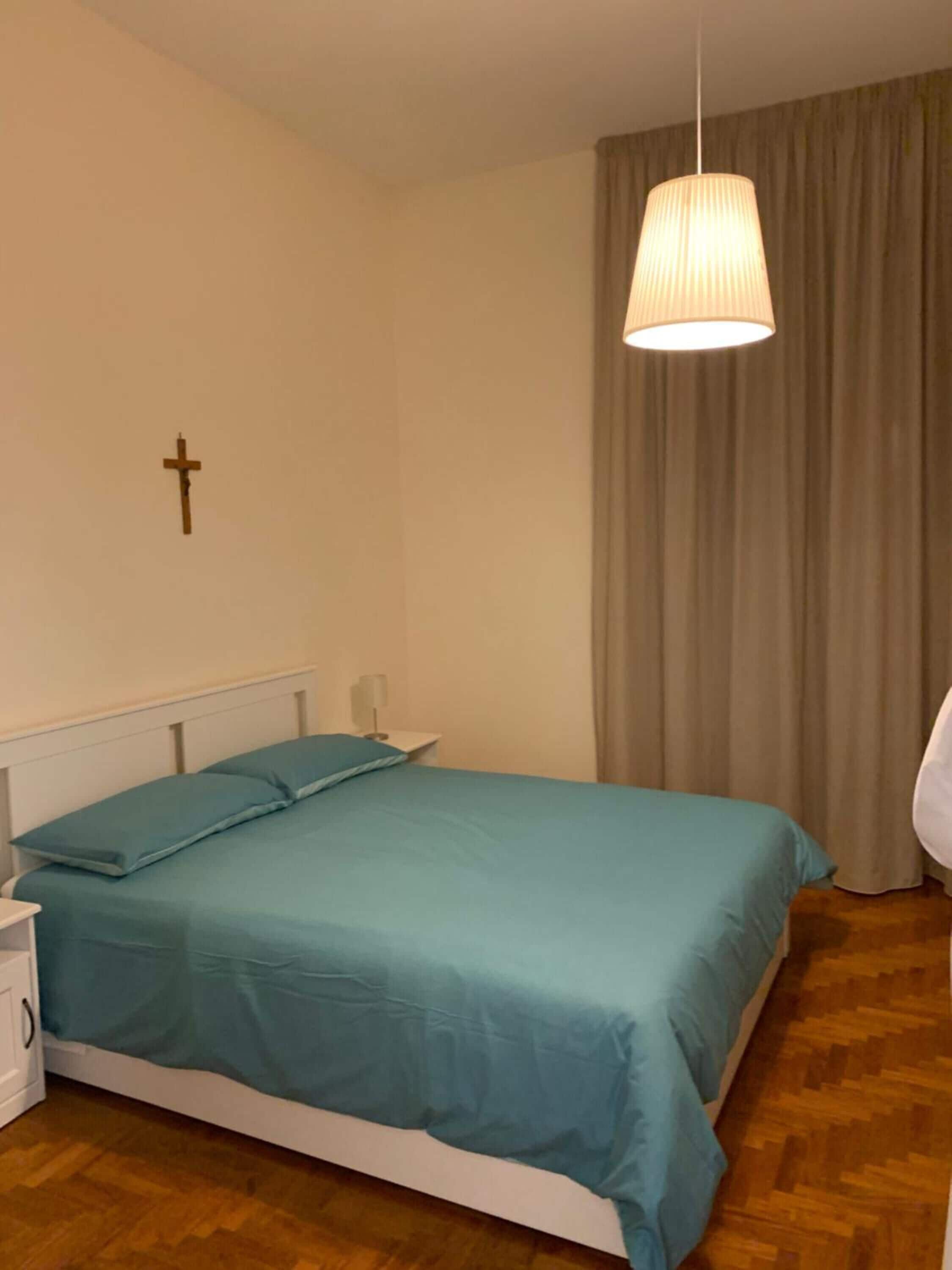 Un dormitorio con cama, una cruz en la pared, una lámpara y una cortina.