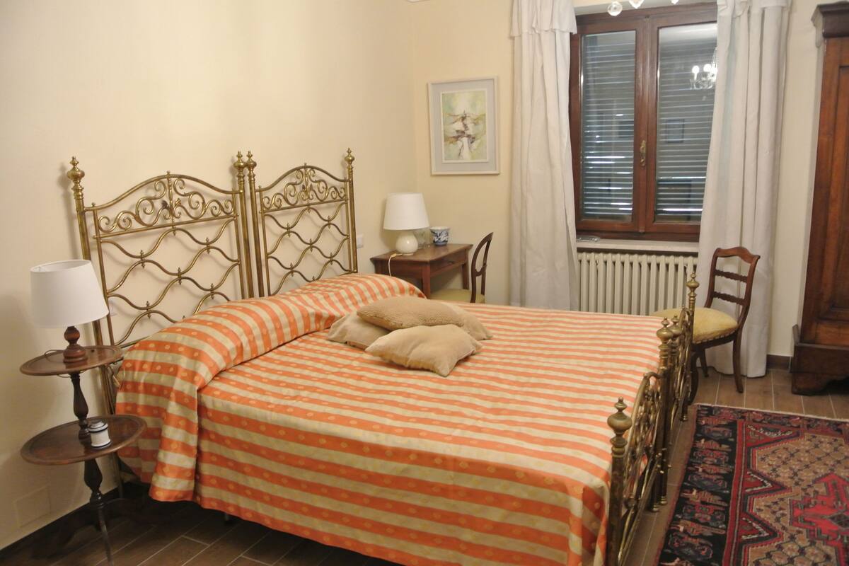 Una cama bien hecha con ropa de cama a rayas, una mesita de noche con una lámpara, una silla y una ventana con cortinas.