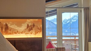 A room with a large window overlooking a snowy mountain landscape, a plaid armchair with a red pillow, and a wall-mounted picture frame. - Naturhotel Waldesruhe (Oberstdorf)