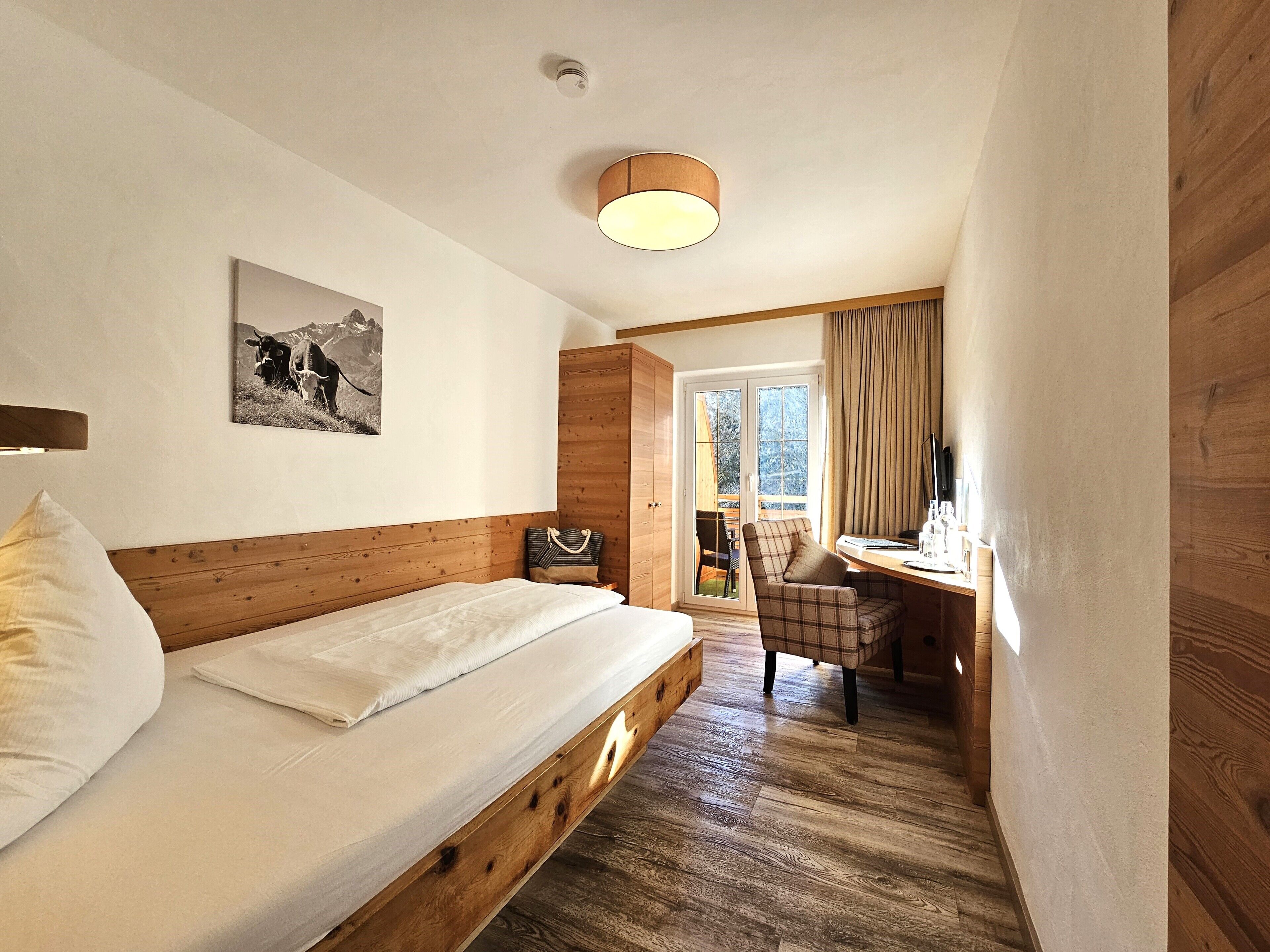 Un dormitorio con cama de madera, cabecera, mesita de noche, una silla, un escritorio, ventana con cortinas e imagen colgada en la pared.