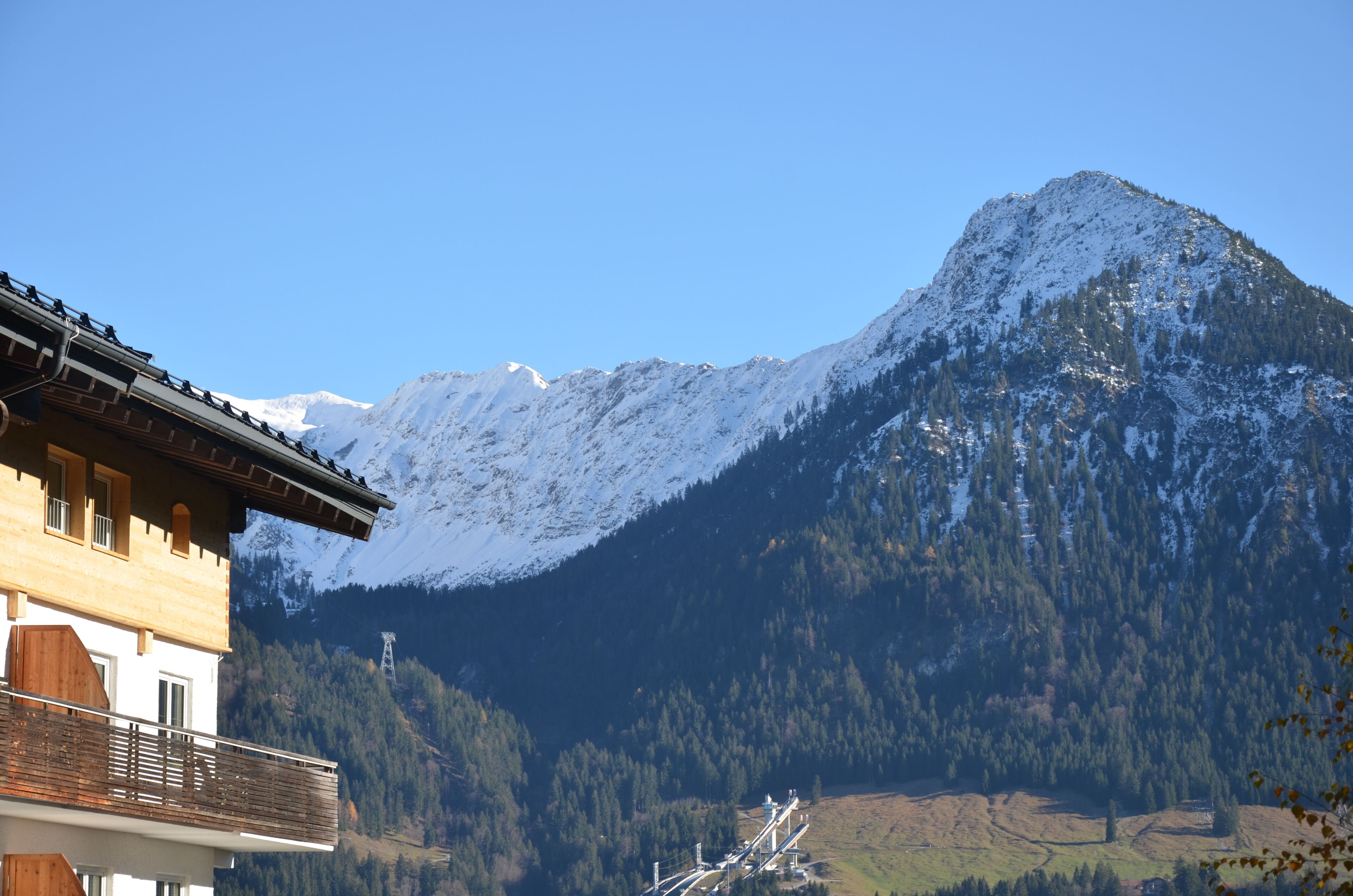 Une montagne aux sommets enneigés, un bâtiment en bois doté d’un balcon et un ciel bleu azur.