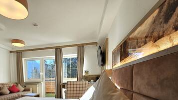 Ein Hotelzimmer mit Bett, Sofa, Esstisch, Fernseher und einem großen Fenster mit Blick auf Bäume und Berge.