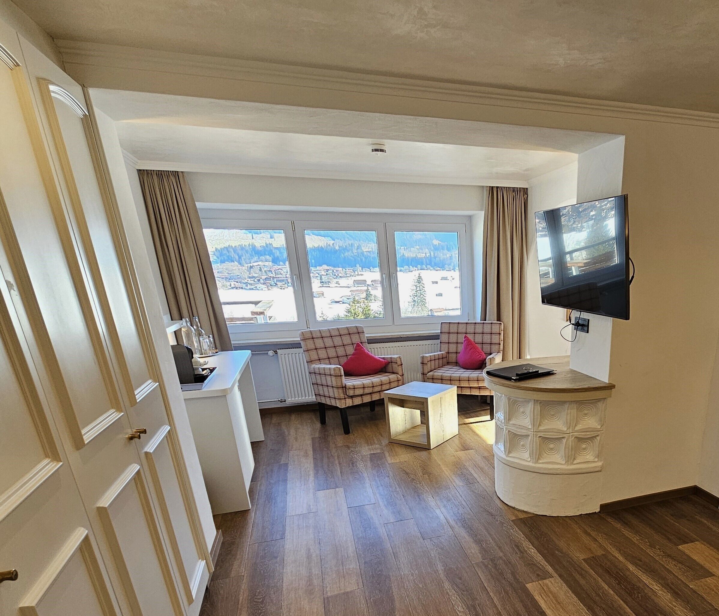 A compact hotel room with a wooden floor, a small desk with a chair, a television mounted on the wall, and a window offering a view of a snowy landscape.