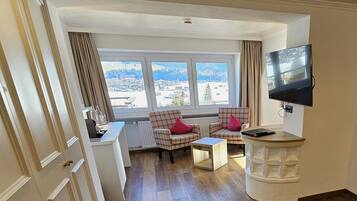 A compact hotel room with a wooden floor, a small desk with a chair, a television mounted on the wall, and a window offering a view of a snowy landscape.