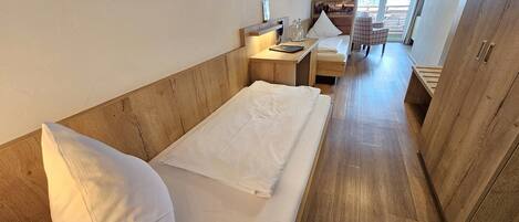 A compact hotel room with a wooden headboard, a single bed, a small desk with a chair, and a view of the outdoors through a window.