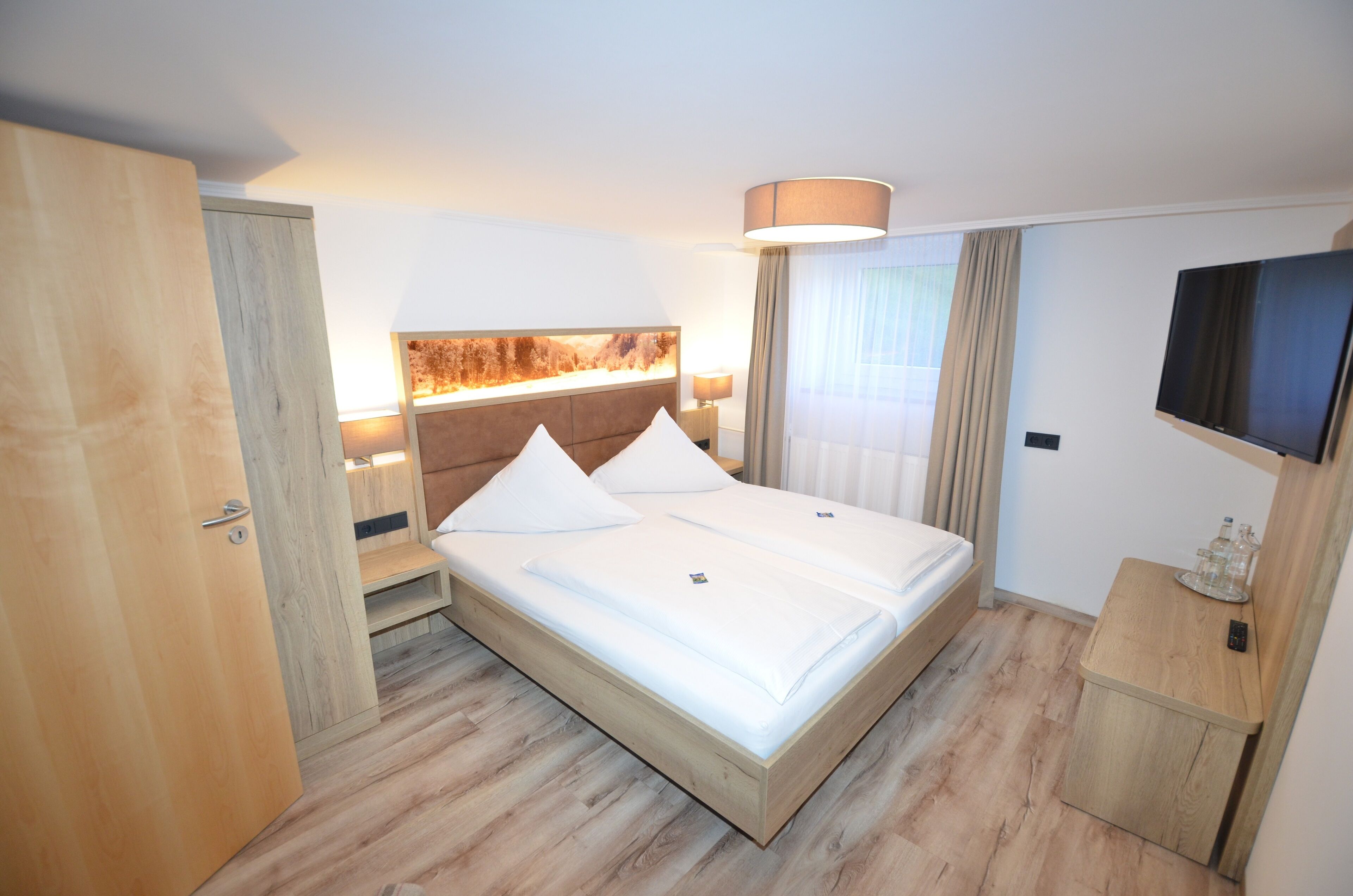 A hotel room with a wooden bed, a flat-screen TV, a bedside table, and a large window with curtains.