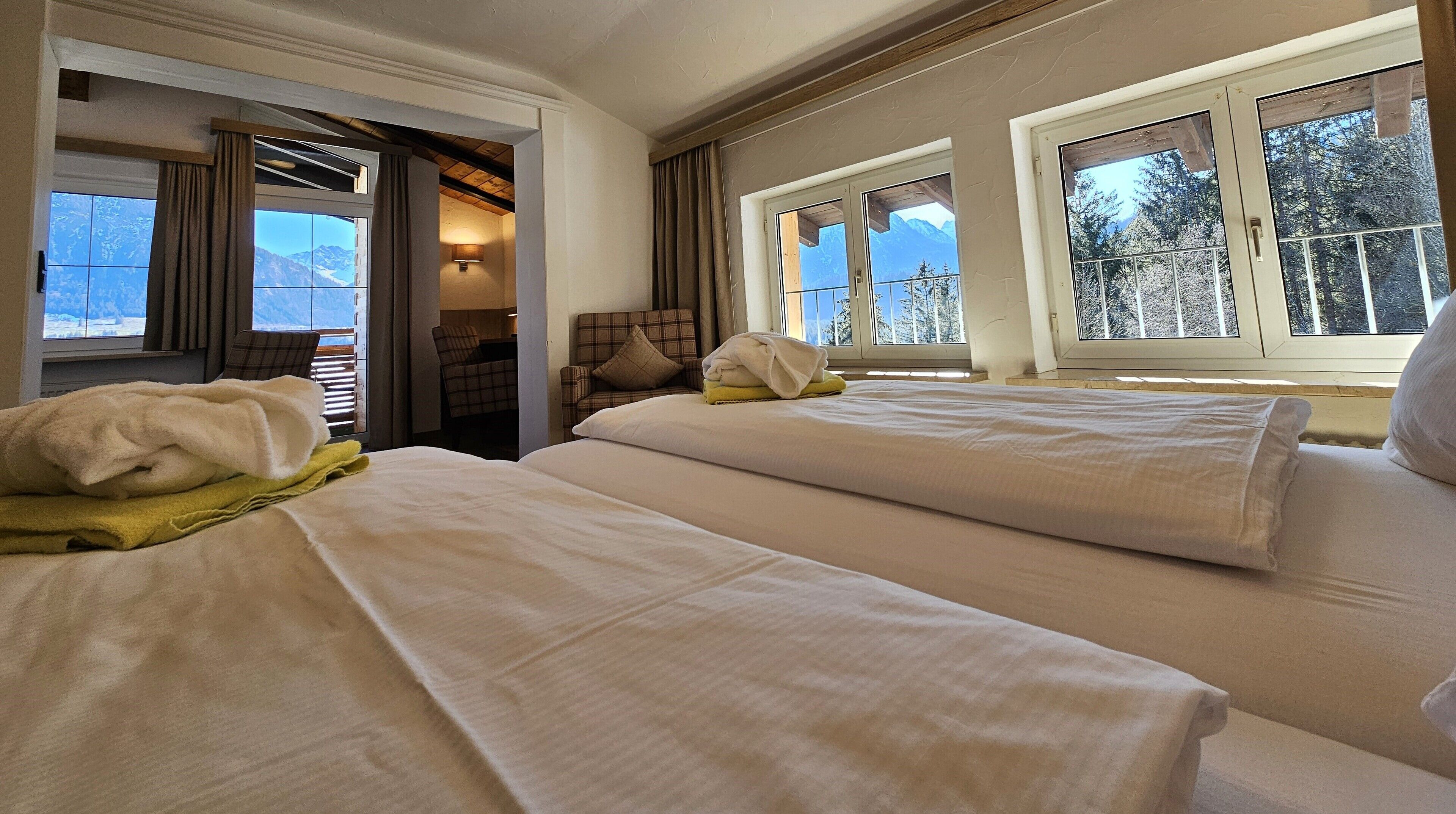 Habitación de hotel con dos camas, vista a montañas a través de amplios ventanales y un rincón de descanso acogedor con una lámpara.