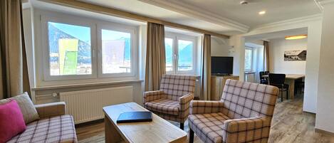 A cozy living room with plaid furniture, a wooden coffee table, and a television. Large windows offer a view of mountains and a hotel sign.