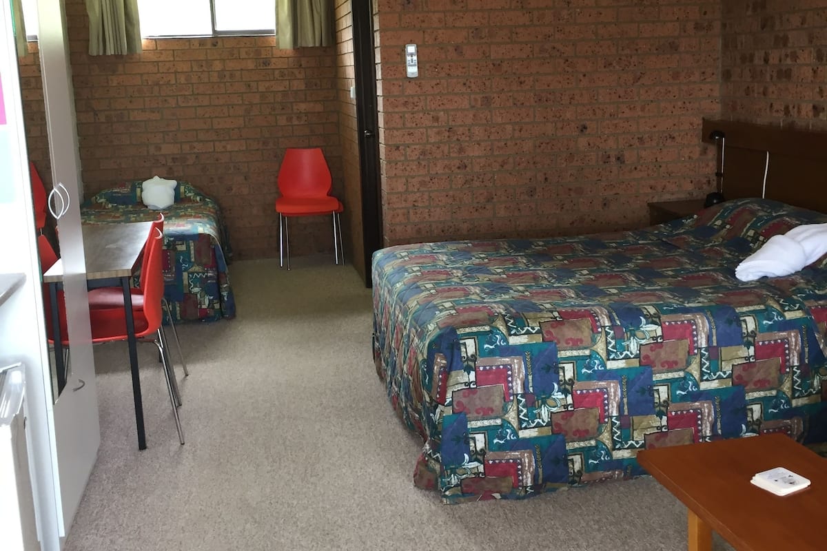 A room with a bed, a desk, a chair, and a table. There is a window with curtains, a ceiling fan, and an air conditioning unit.