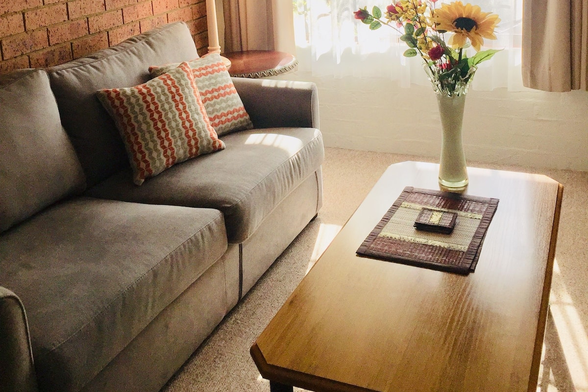 A living room with a grey sofa, a wooden coffee table, a vase with flowers, a lamp, and a brick wall with a framed picture.