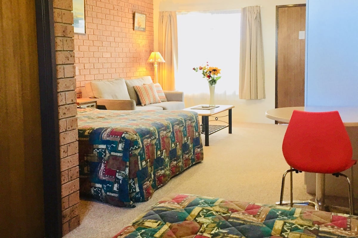 A hotel room with a brick accent wall, a bed with a colorful quilt, a red chair, a small table with a lamp, and a view of the outdoors.
