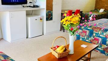 Una habitación acogedora con una televisión mostrando 'Officeworks' en la pared, un refrigerador pequeño, una mesa de madera con una cesta de fruta, un florero y una colcha de patchwork colorida.
