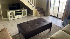 A living room with a staircase, a television on a cabinet, a brown leather ottoman, and a sliding glass door to the outside. - SUNDIAL - SITTING IN THE SAND. 3 BR, 2 BA. POOL. LESS CROWDED AREA OF BEACH. (Gulf Shores)