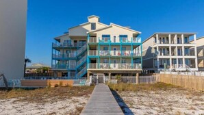 A beachfront hotel with multiple balconies, a wooden boardwalk leading to the entrance, and a clear blue sky. - SUNDIAL - SITTING IN THE SAND. 3 BR, 2 BA. POOL. LESS CROWDED AREA OF BEACH. (Gulf Shores)