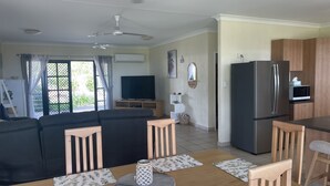 Ein modernes Wohnzimmer mit einer schwarzen Couch, Holzstühlen, einem Esstisch mit Platzdeckchen, einem Fernseher und einer Küchenzeile inklusive Kühlschrank und Mikrowelle.
