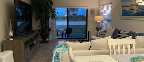 A living room with a TV, a large potted plant, a sofa, a table, and a view of a body of water through a sliding glass door.