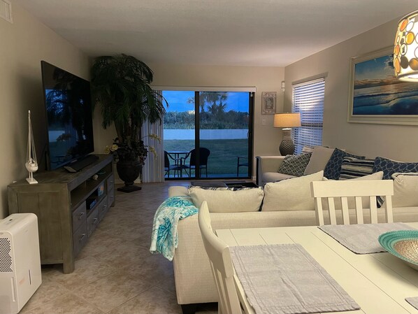 A living room with a TV, a large potted plant, a sofa, a table, and a view of a body of water through a sliding glass door. - Incredible three bedroom oceanfront corner unit. Out to the beach in seconds! (Cocoa Beach)