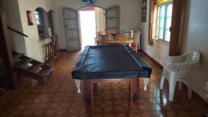 Een kamer met een pooltafel, houten stoelen, een trap en een raam met gordijnen.