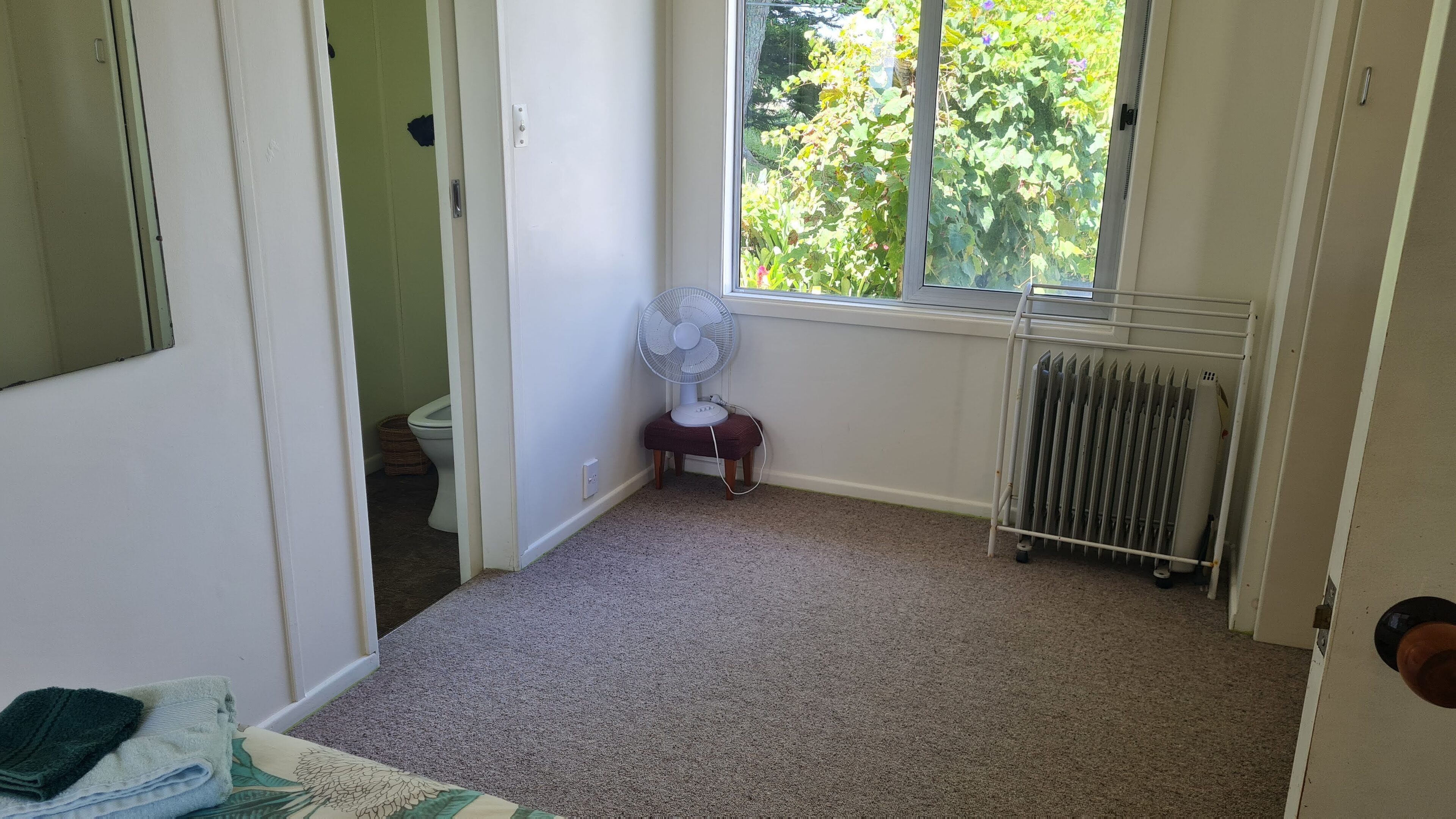 A room with a radiator, a window with a view of greenery, a fan, and a corner with a toilet and a basket.