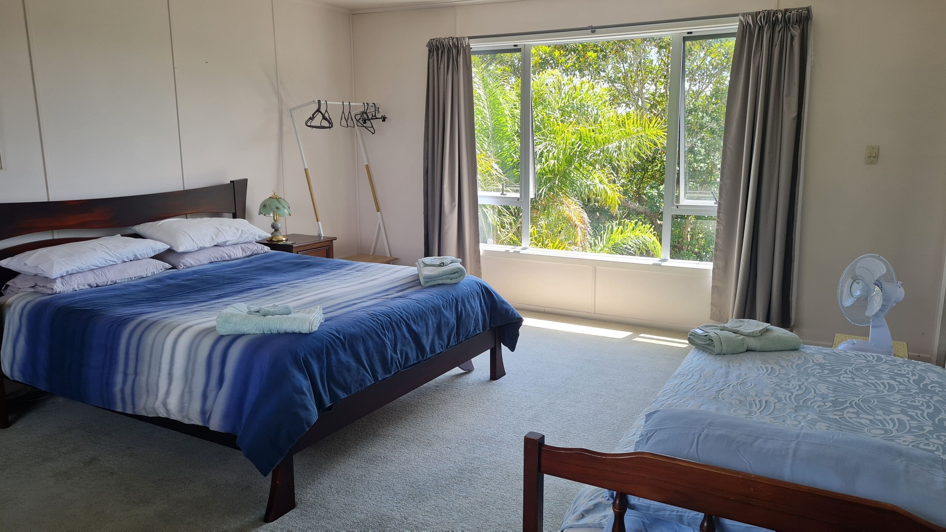 A bedroom with a large bed, a smaller bed, a fan, a window with curtains, and a wall-mounted drying rack.