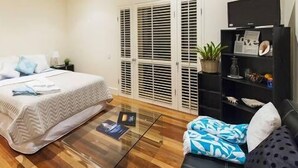 A bedroom with a bed, bedside tables, a glass coffee table, a sofa, a TV, and a window with blinds. - "Riverbend Suites" BYRON Hinterland Retreat (Rosebank)