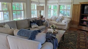 A spacious living room with a large beige sectional sofa, a smaller white sofa, and a coffee table. There are multiple windows with white blinds, a wooden cabinet with a TV, and a patterned rug on the floor. - Stunning hse-AC, heatd Pool, spa, fire pit, private acre, walk/free bus to town (Nantucket)