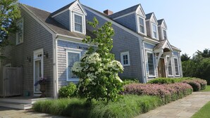 A two-story house with a grey siding exterior, a front porch, and a well-maintained garden with various plants and flowers. - Stunning hse-AC, heatd Pool, spa, fire pit, private acre, walk/free bus to town (Nantucket)