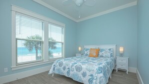 Un dormitorio con cama, dos mesitas de noche, ventilador de techo y vistas a la playa a través de las ventanas.