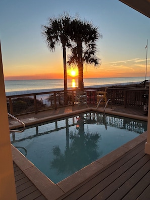 Un área de piscina con vista al atardecer sobre el océano, terraza de madera y una palmera.