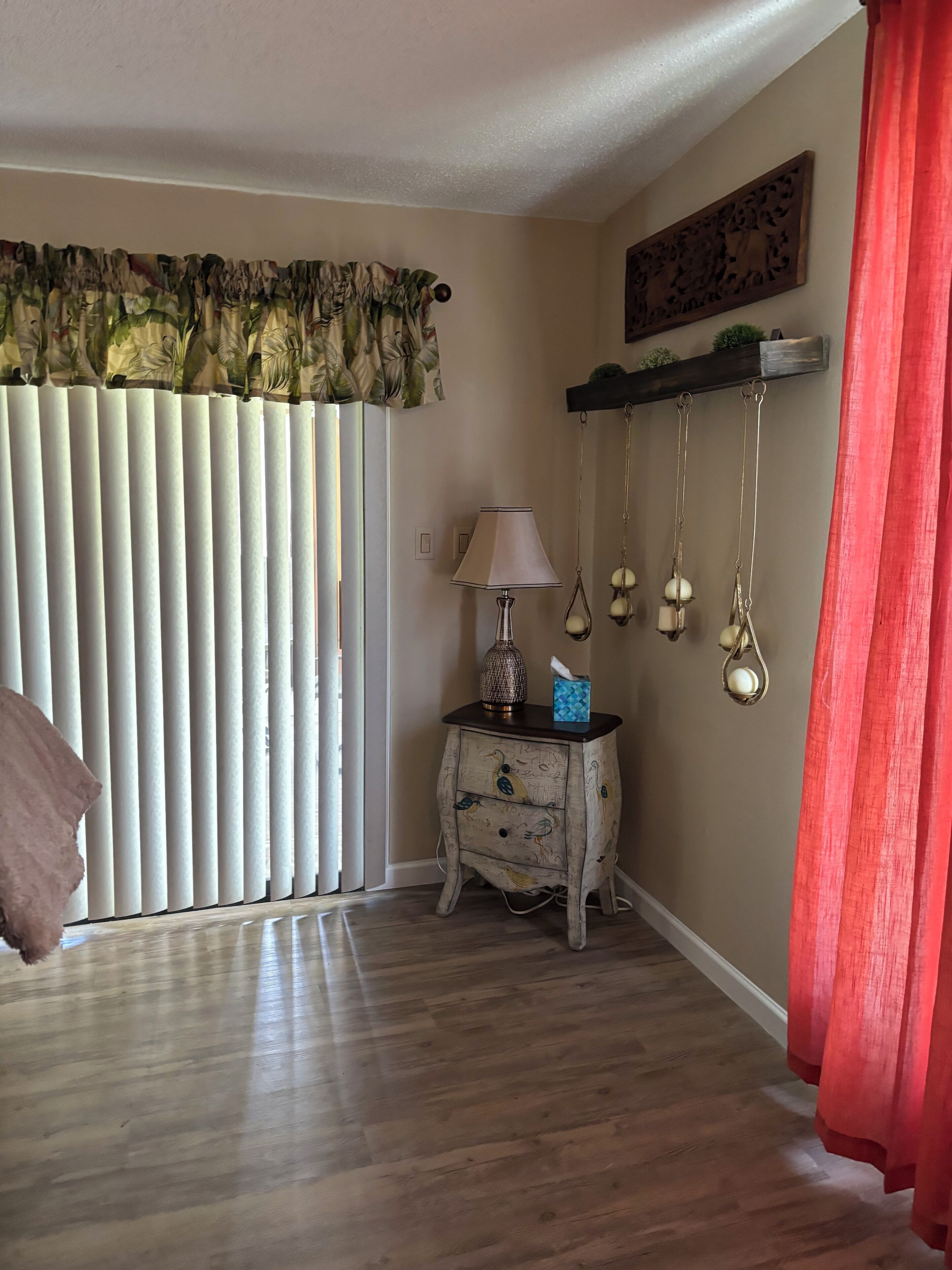 A room with a wooden floor, a white-framed window with vertical blinds, a small wooden nightstand with a lamp and decorative items, and a red curtain.
