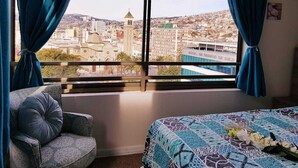 Ein Hotelzimmer mit großem Fenster und Blick auf die Stadt, einem Bett mit gemustertem Überwurf und einem Sessel mit Kissen.
