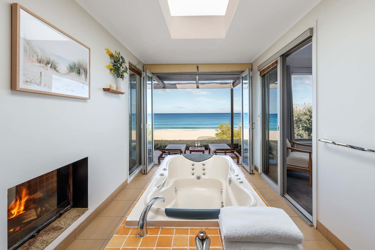 A modern bathroom with a large bathtub, a fireplace, and a view of the beach.