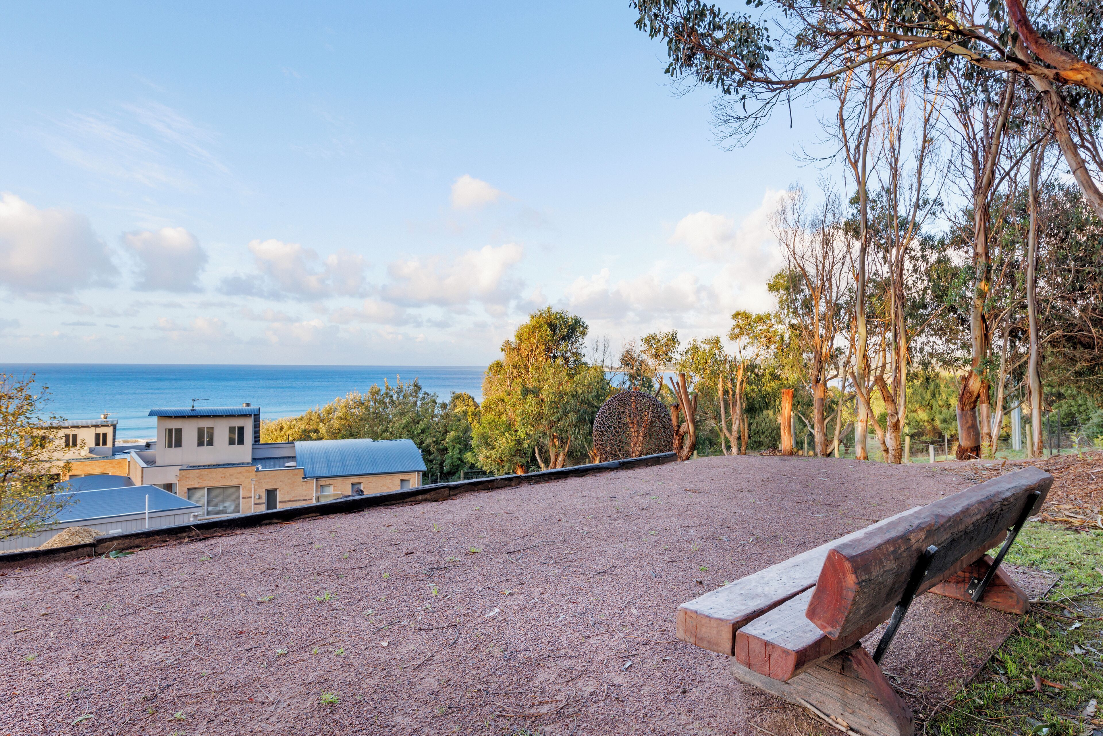 A park with a wooden bench, a dirt path, and a view of buildings and the sea.