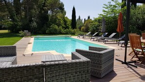 Une piscine entourée d’une terrasse en bois, agréée de chaises longues et de meubles en osier, le tout baigné de verdure.