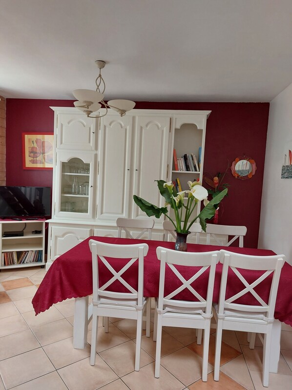 Un comedor con mantel rojo, sillas blancas, un armario blanco con puertas de cristal y un televisor sobre un mueble.