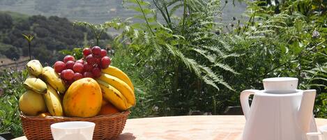 A table with a fruit basket, a teapot, and cups on a tiled patio with a mountainous backdrop.