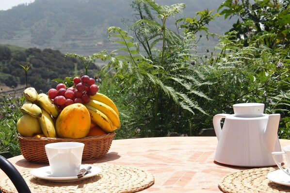 A table with a fruit basket, a teapot, and cups on a tiled patio with a mountainous backdrop.