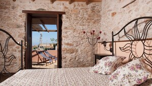 A bedroom with a stone wall, a bed with floral bedding, a candelabra, and a view of a terrace with chairs and a table.