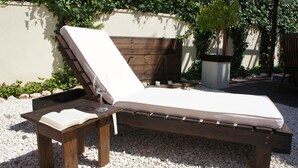Una silla de lounge de madera con un cojín blanco, una mesita de madera con un libro abierto y una planta en maceta.