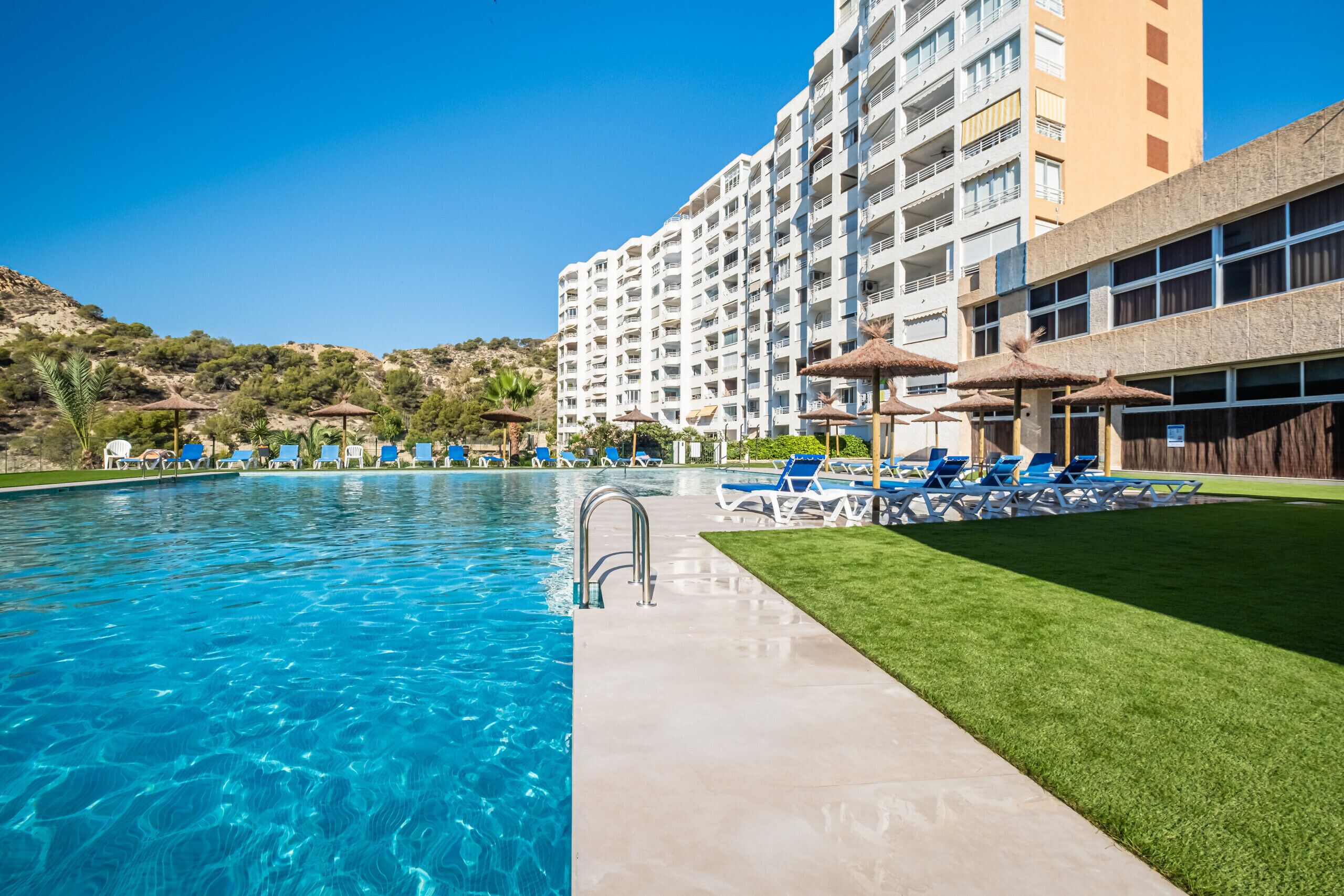 Une piscine à l’eau d’un bleu cristallin, entourée de chaises longues et de parasols, avec un bâtiment d’hôtel en arrière-plan.
