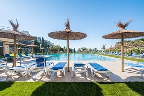 A poolside area with lounge chairs and thatched umbrellas, surrounded by greenery and buildings in the background. - Eurotennis 405-1 (Villajoyosa)