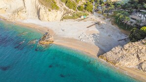 Luchtbeeld van een strand met helder turquoise water, rotskliffen en een klein gebouw met een wit dak.